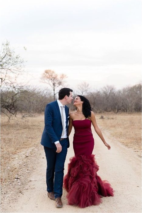 Una pareja de novios camina por un paraje de campo amarillento, ella viste vestido rojo quemado