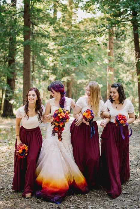 Una novia con vestido blanco y de colores naranja, rojo y amarillo con sus damas vestidas de blanco y rojo quemado caminan entre árboles