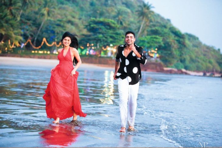 una pareja va corriendo por la playa, él con camisa negra con blanco y pantalón blanco y ella con vestido rojo