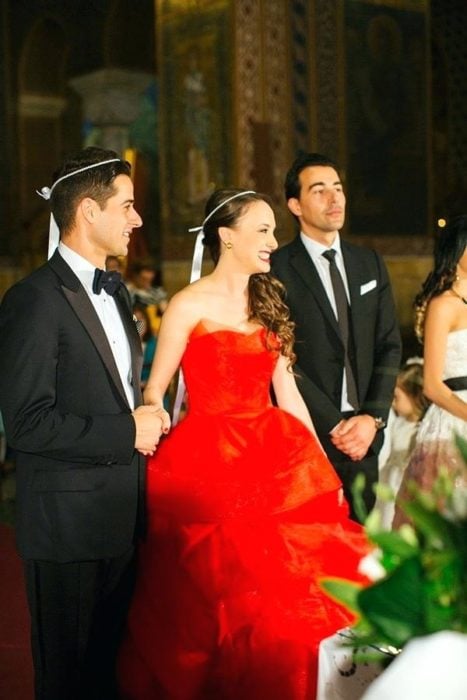 Una pareja de novios y su padrino frente al altar, ella viste vestido rojo estrapless y moño blanco en la cabeza