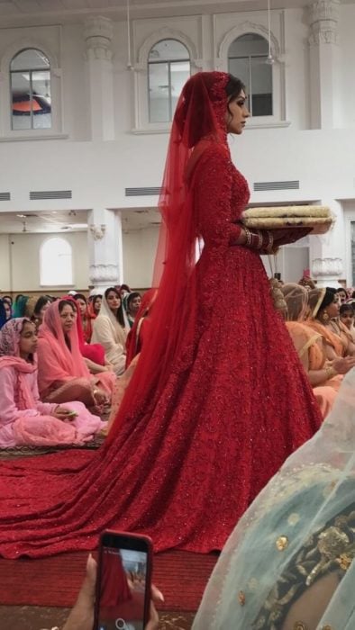 Una novia hindú con vestido rojo camina hacia el altar en un templo lleno de personas sentadas en el piso