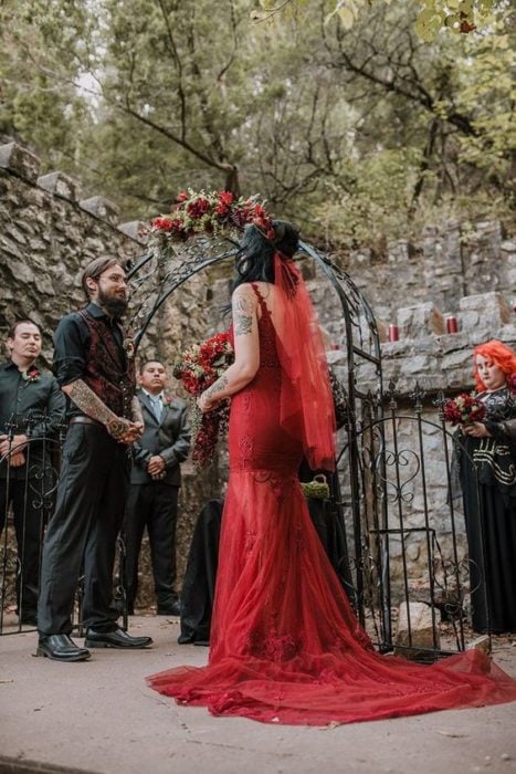una pareja de novios se casa en un jardín, ella con vestido rojo entallado y velo rojo
