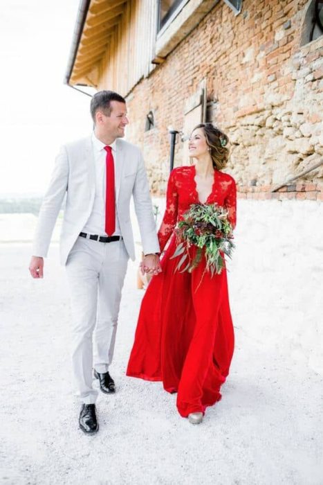 Una pareja tomada de la mano camina a un lado de un edificio de piedra, él con traje gris y corbata roja y ella vestido rojo y con su ramo
