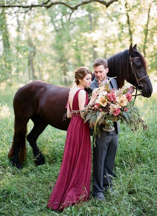 Una pareja de novios posa a un lado de un caballo frisón negro, ella con vestido rojo quemado y el novio sostiene el ramo