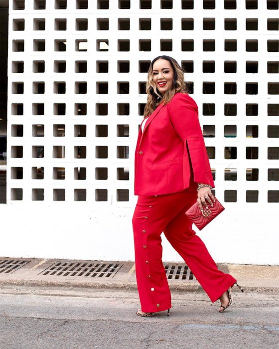 una mujer camina frente a una barda de cuadros, va vestida con traje de pantalón y saco rojos y un bolso de mano rojo