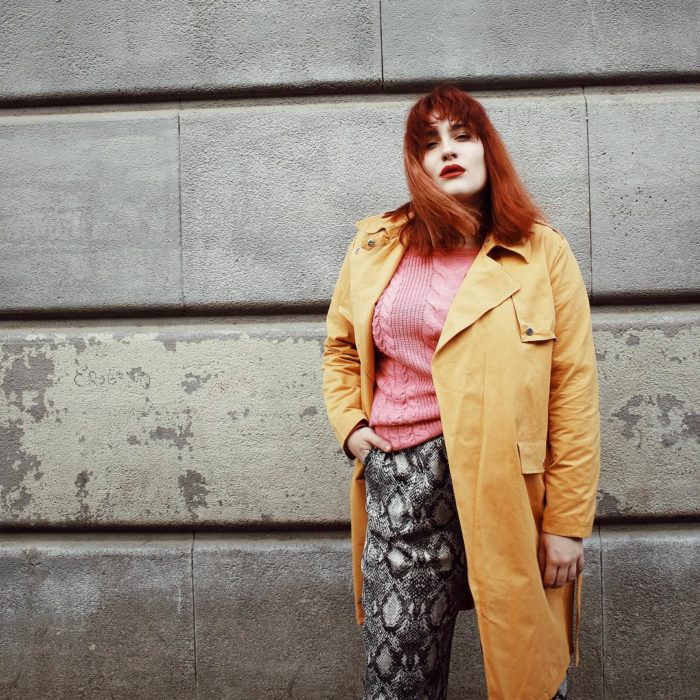 Una mujer posa frente a una pared de concreto con un pantalón de animal print, un suéter rosa y una gabardina amarilla