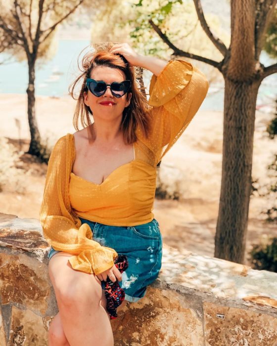 una mujer sentada en una bardita en un paraje natural, vestida con short de mezclilla y blusa mostaza y lentes de corazón