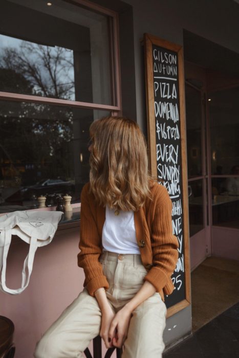 Outfits con cardigans; chica en cafetería con cabello castaño rojizo, suéter anaranjado con botones y pantalón caqui