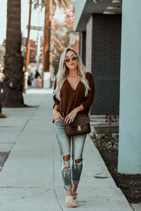 Outfits con cardigans; chica de cabello rubio platinado caminando por la calle, vestida con suéter de botones café y pantalón desgastado de mezclilla