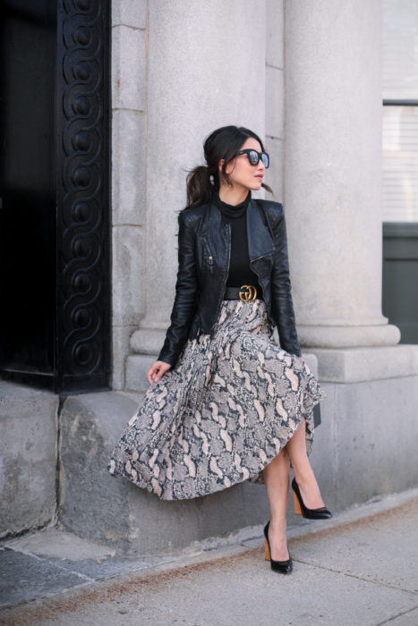 Mujer sentada afuera de edificio usando lentes grandes de sol, chaqueta de cuero negra y falda larga de animal print de serpiente