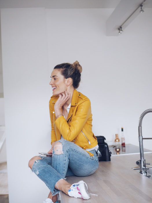 Chica sentada en barra en la cocina, con peinado de chongo, vistiendo chaqueta de piel amarilla y pantalón de mezclilla desgastado