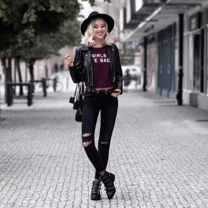 Chica rubia posando para foto en la calle, con sombrero boho, chamarra de cuero, blusa básica guinda, jeans entubados y botas con estoperoles; con cámara analógica 