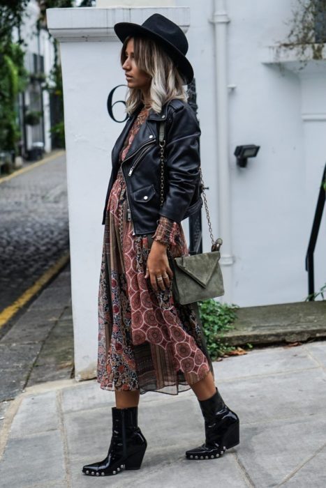 Chica con estilo boho, sombrero, chaqueta de cuero negro, vestido hippie y botas de tacón grueso con estoperoles 