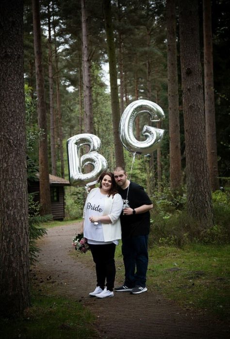 Rebecca y Glen Maxwell en un bosque con una cabaña de fondo y un globo plateado cada quien con la inicial de su nombre