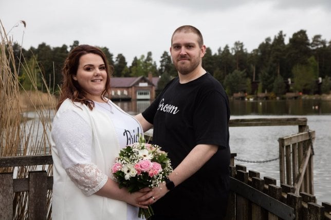 Rebecca y Glen Maxwell posan con el ramo de novia a la orilla de un lago, al fondo se ve un bosque y una cabaña de madera