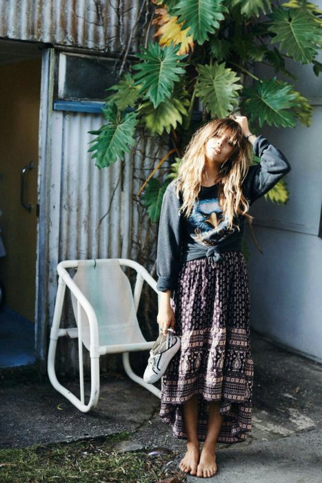 Ropa estilo boho o hippie chic; mujer parada frente una planta, de cabello castaño largo, con sudadera oversized, maxi con los tenis blancos en las manosfalda