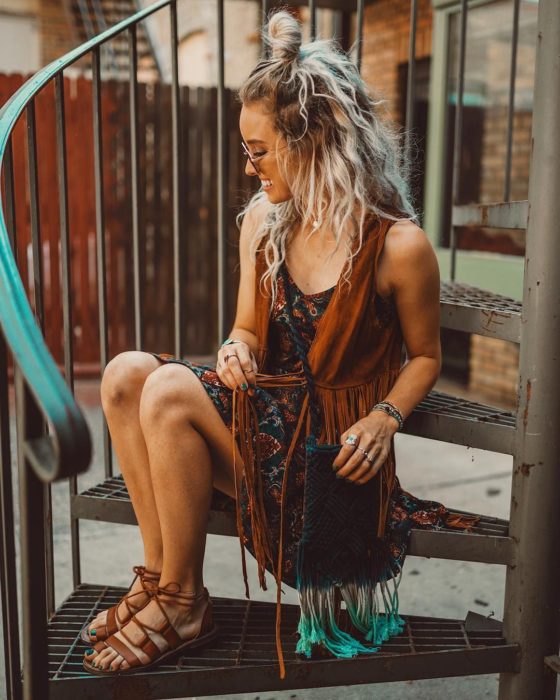 Ropa estilo boho o hippie chic; chica sentada en escaleras de caracol, con vestido, chaleco de flecos café con sandalias de piso