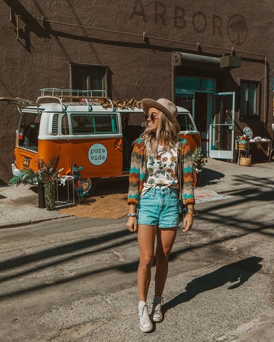 Ropa estilo boho o hippie chic; chica frente a una van, sonriendo con sombrero, lentes de sol, playera con estampado de flores, abrigo tejido de colores, short de mezclilla y tenis blancos converse