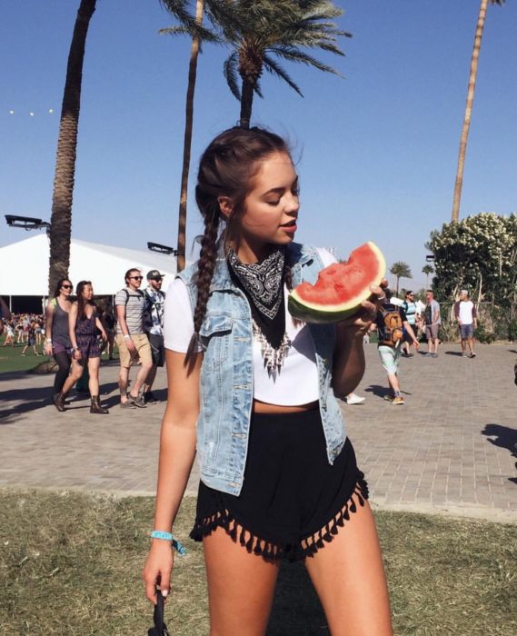 Ropa estilo boho o hippie chic; chica con peinado de trenzas de boxeadora con crop top, chaleco de mezclilla, short con flecos, paliacate negro en el cuello, comiendo una rebanada de sandía