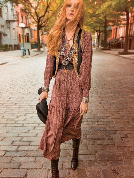 Ropa estilo boho o hippie chic; mujer de cabello largo y pelirrojo en calle empedrada con vestido de flores, cinto, con chaleco, pulseras y collares