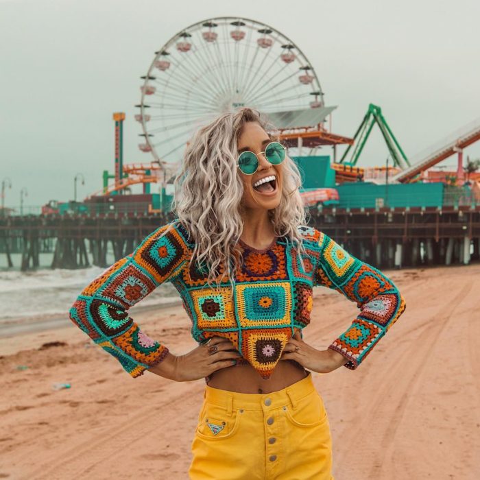 Ropa estilo boho o hippie chic; chica de cabello rubio platinado chino hasta los hombros, con crop top tejido, con jean amarillo, sonriendo con las manos en la cintura frente a un parque de diversiones con rueda de la fortuna