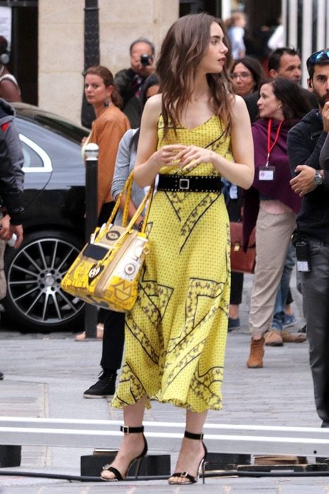 Lily Collins caminando por las calles de París mientras usa un vestido de color amarillo con un cinturón de gamuza color negro 