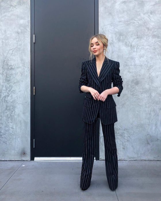 Famosas con traje; Sabrina Carpenter con saco y pantalón de vestid negro con rayas blancas verticales