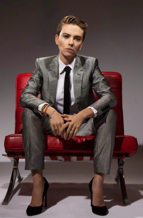 Famosas con traje; Scarlett Johansson con cabello corto pixie, con saco y pantalón de vestir gris briññante, con camisa blanca y corbata negra, sentada en un sillón rojo