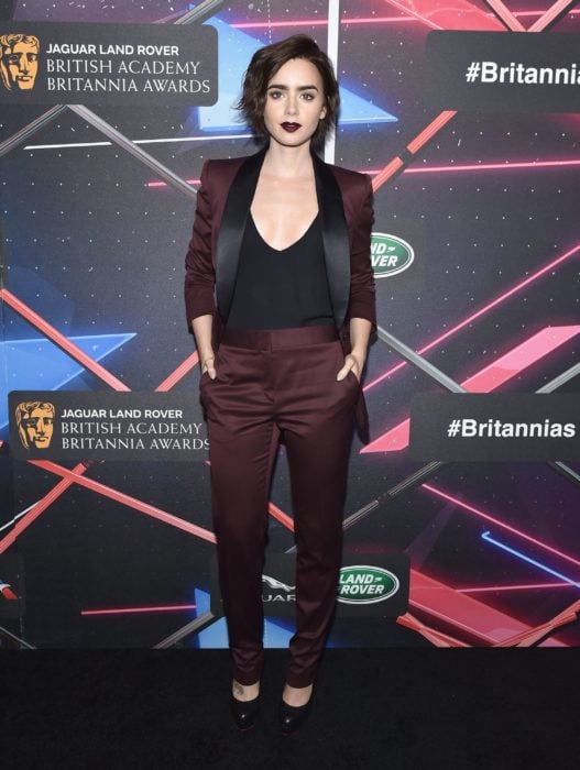 Famosas con traje; Lily Collins en los British Academy Britannia Awards con saco y pantalón de vestir color rojo vino y camisa negra, con cabello corte pixie