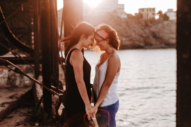 Pareja de chicas tomadas de la mano y uniendo sus frentes mientras están en una puesta de sol 