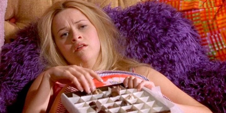 una mujer con cara de enferma comiendo chocolates acostada en la cama