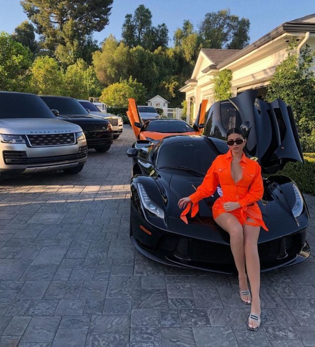Kylie Jenner sobre un coche deportivo con las puertas abiertas, afuera de su casa con muchos coches más
