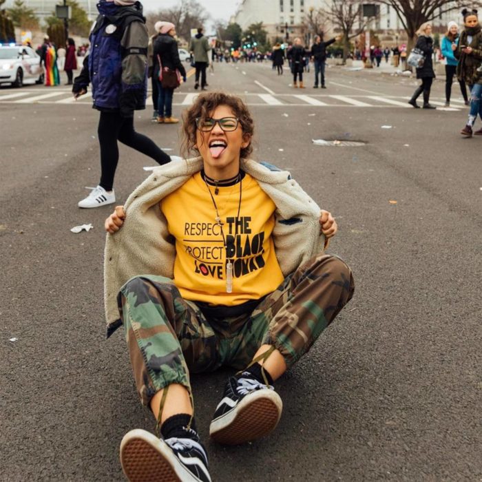 Looks tomboys; chica con atuendo masculino, Zendaya sentada en la calle con pantalón militar, playera amarilla y chamarra de mezclilla con borrega, sacando la lengua