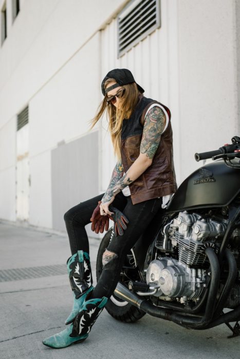 Looks tomboys; chica con atuendo masculino con chaleco y botas vaqueras, sentada en motocicleta estacionada