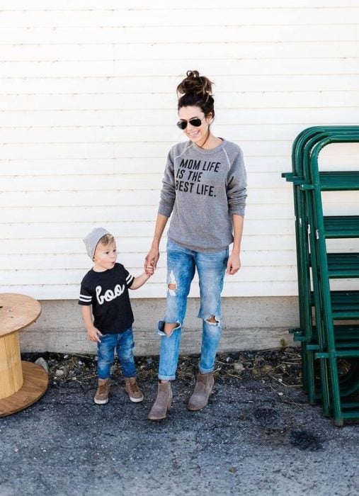 Madre e hijo tomados de la mano, compartiendo outfits con jeans rotos, sudaderas ligeras y botas en gris