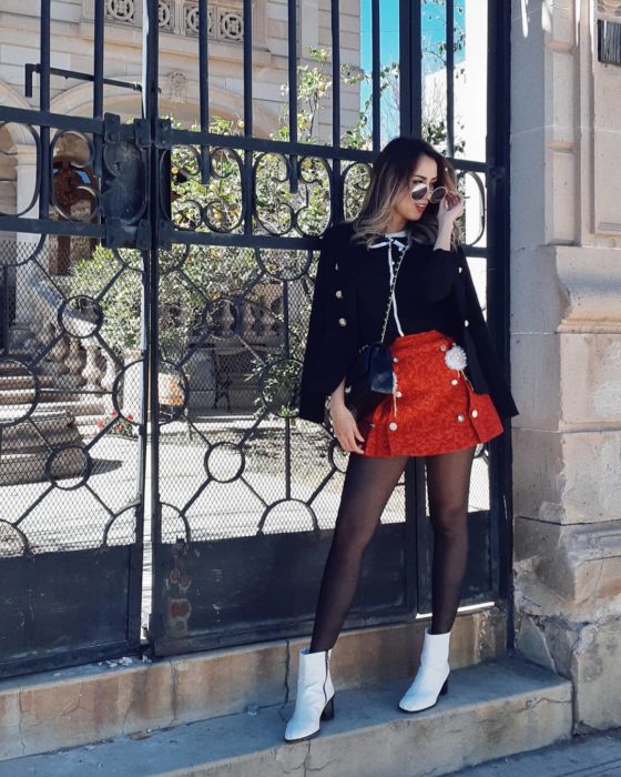 Chica parada frente a una puerta posando para una foto mientras usa una blusa negra, falda de color rojo y botines blancos 