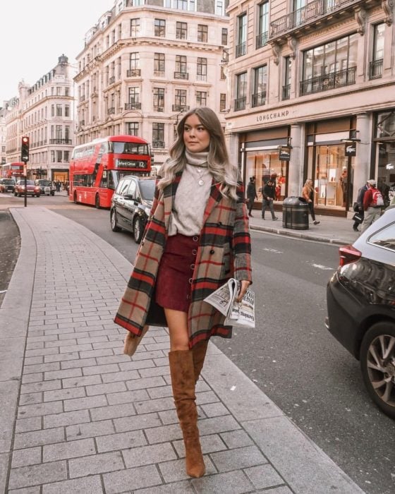 Outfits para otoño; chica de cabello platinado caminando en la calle con suéter de cuello de tortuga, abrigo de cuadros, falda de pana y botas cafés largas