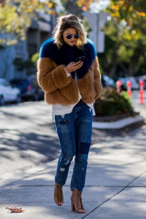 Outfits para otoño; mujer mirando su celular, con abrigo afelpado azul y café, con mom jeans y lentes grandes y redondos