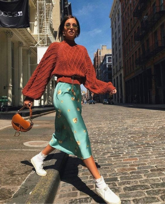 Outfits para otoño; chica cruzando la calle empedrada, con suéter oversized de abuelita, color anaranjado y falda de satín verde menta floreada, con tenis blancos