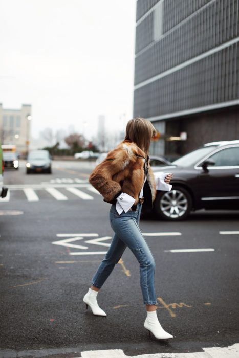 Outfits para otoño; mujer cruzando la calle con abrigo peludo café, jeans azules y botines blancos