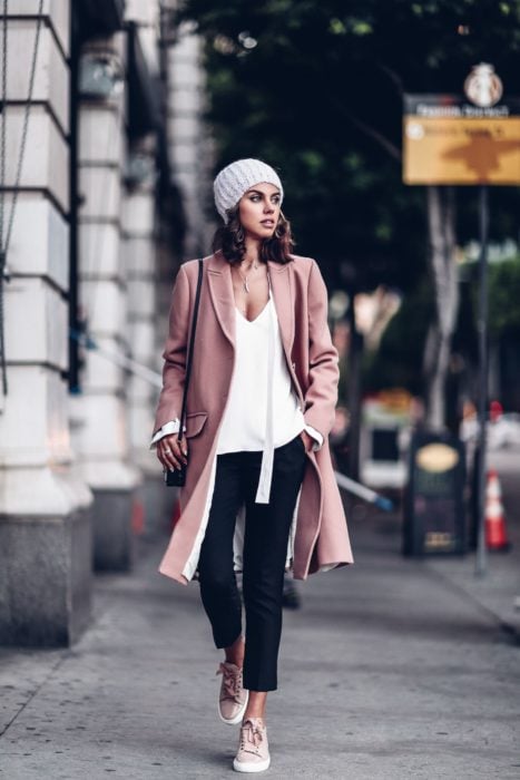 Chica caminando por la calle mientras usa unos jeans negros, blusa blanca, boina y saco rosa 