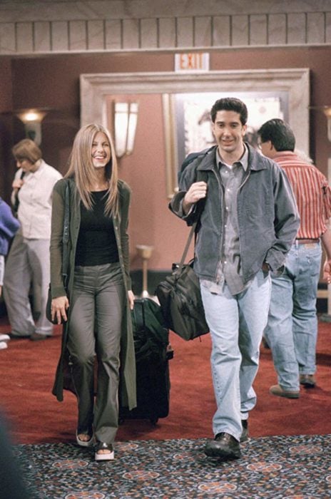 Escena de Friends con Rachel y Ross caminando por un pasillo con maletas, ella viste de gris con negro y él mezclilla con gris