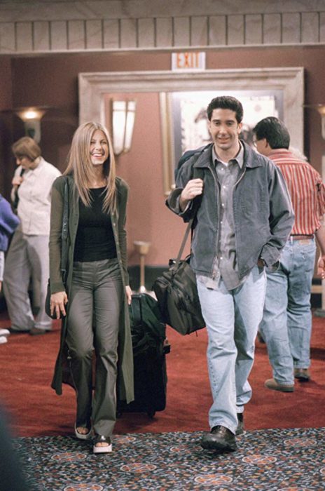 Escena de Friends con Rachel y Ross caminando por un pasillo con maletas, ella viste de gris con negro y él mezclilla con gris