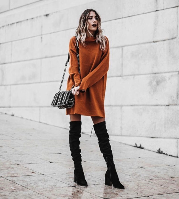 Chica de cabello rubio usando suéter oversized anaranjado como vestido de otoño con botas negras largas de tacón cuadrado 