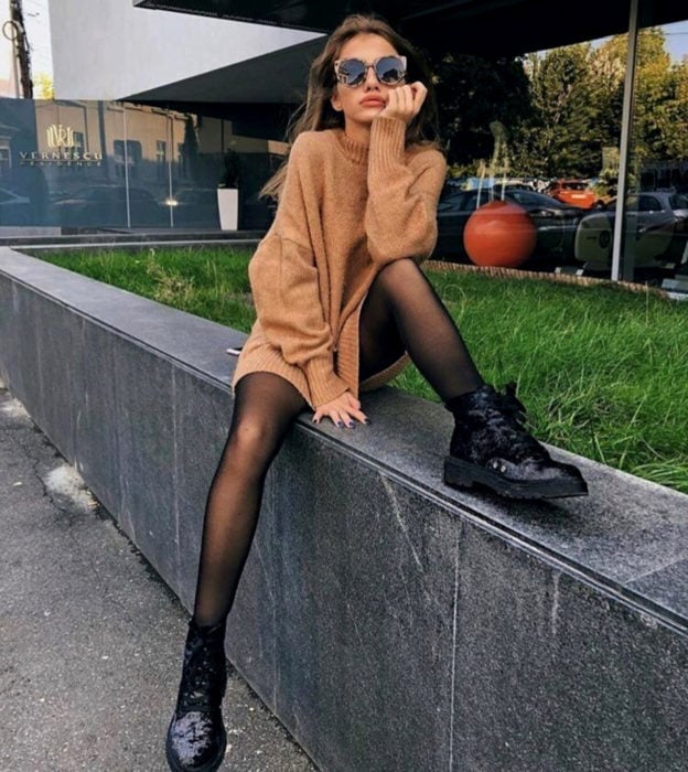 Chica sentada en la calle con lentes de sol, suéter largo como vestido de otoño, medias negras y botas de trabajo Dr. Martens brillantes
