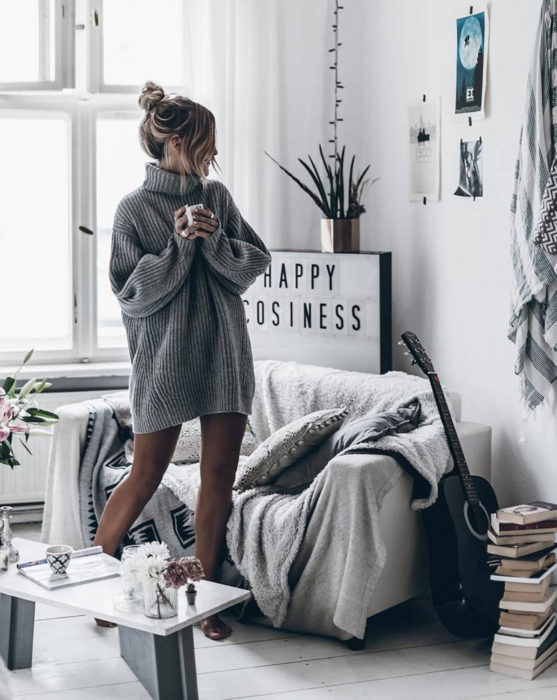 Mujer con peinado de chongo despeinado, usando suéter gris oversized como vestido de otoño, bebiendo una taza de café en su casa junto a su sillón con guitarra acústica negra y una pila de libros