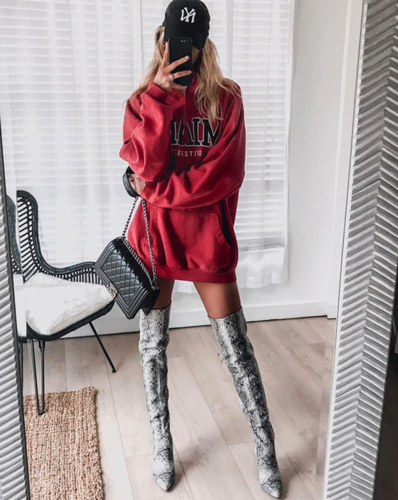 Mujer rubia tomándose selfie frente al espejo, con sudadera roja oversized como vestido de otoño, con botas largas de animal print de serpiente gris