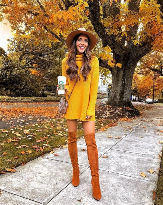 Chica en la calle frente a un árbol de hojas amarillas, usando un sombrero, botas largas anaranjadas y suéter holgado como vestido de otoño