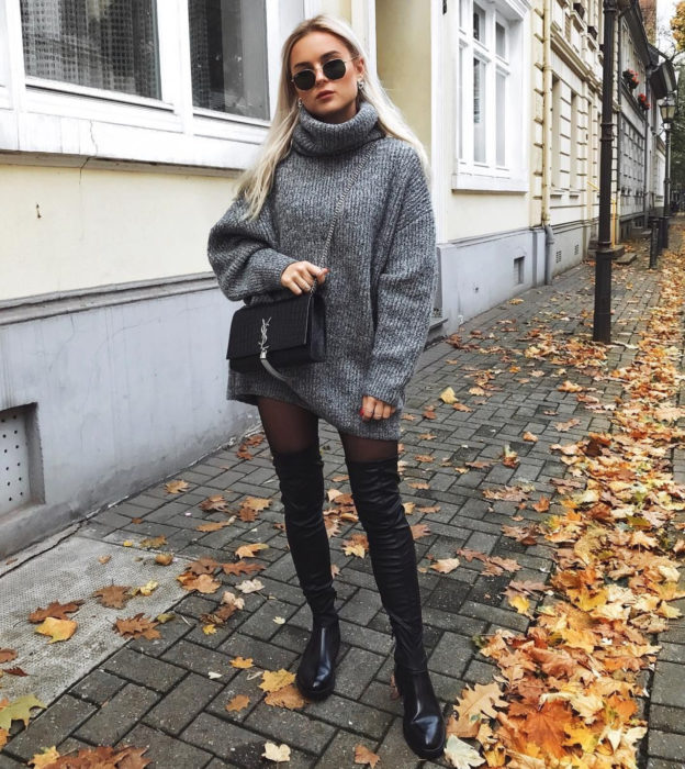Chcia de cabello rubio y largo caminando en la calle con hojas en el suelo, vistiendo un suéter gris y grande como vestido de otoño con medias y botas largas y negras