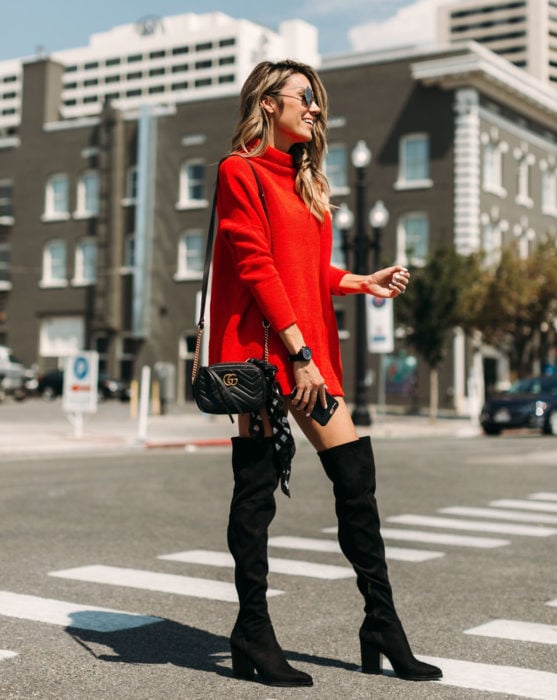 Mujer rubia en la calle, sonriendo y vistiendo un suéter oversized rojo como vestido de otoño con botas negras y largas de gamuza